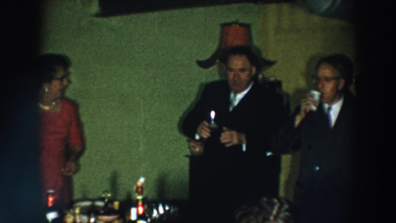 Two men in suits and a woman in a red dress stand with drinks near a table.
