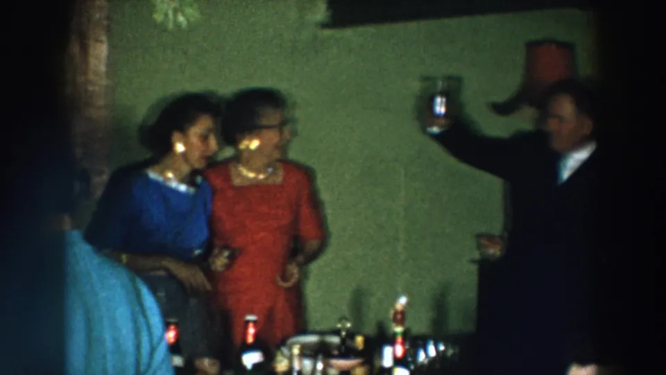 A man raises a glass in a toast as women in formal dresses stand nearby in a dim 1957 Wheeling, Ohio interior.