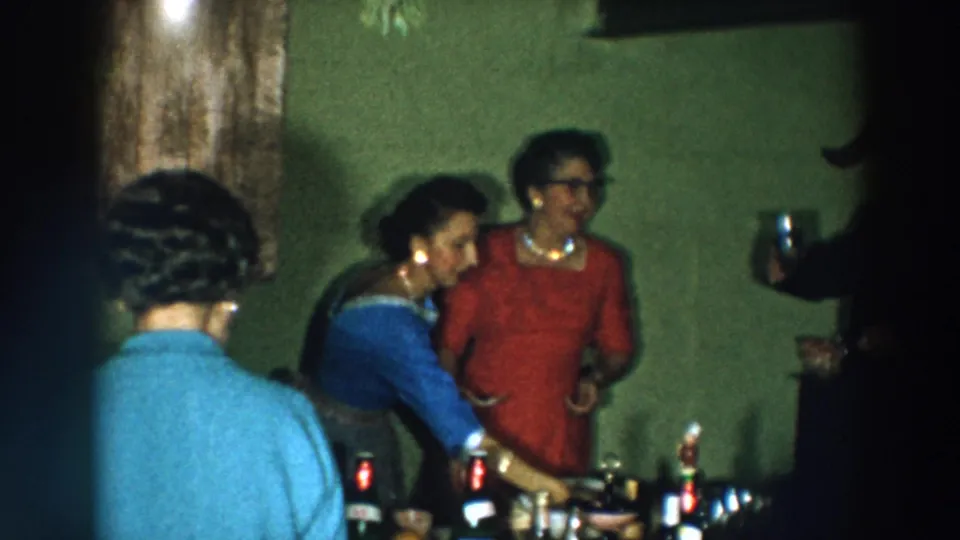 Adults continue the toast in a private 1957 Wheeling, Ohio room, with bottles on the table and a raised glass at frame edge.