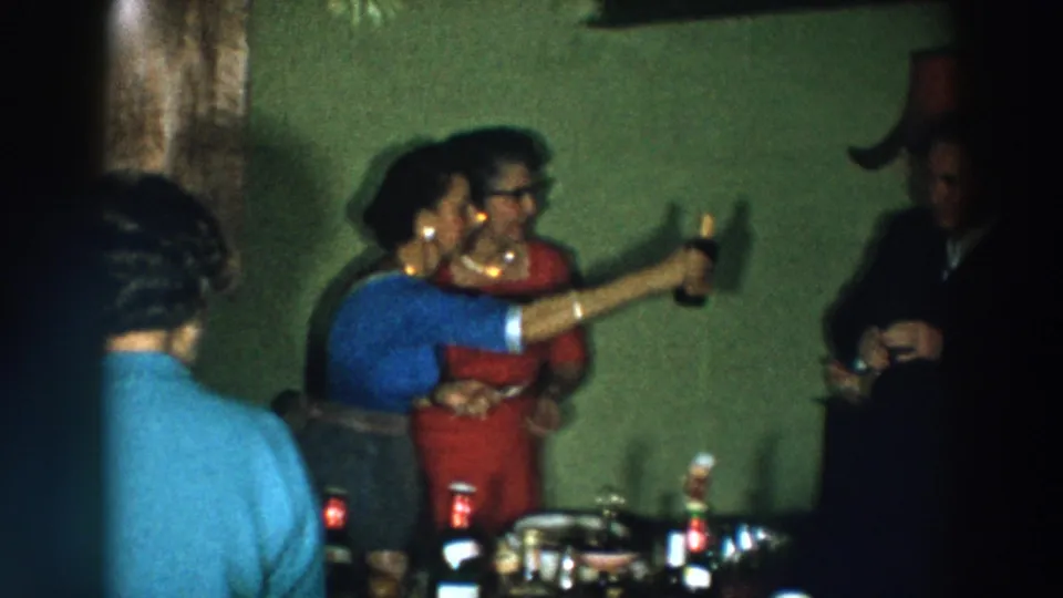 1957 Wheeling, Ohio home-movie frame showing party guests in suits and dresses raising drinks near a table.