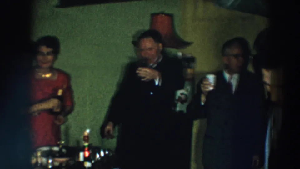 Adults at a 1957 Wheeling, Ohio indoor party stand near bottles and lamps while holding drinks.