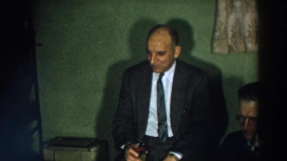 1957 Wheeling, Ohio home-movie frame with a seated man in a suit holding a drink in a private room.