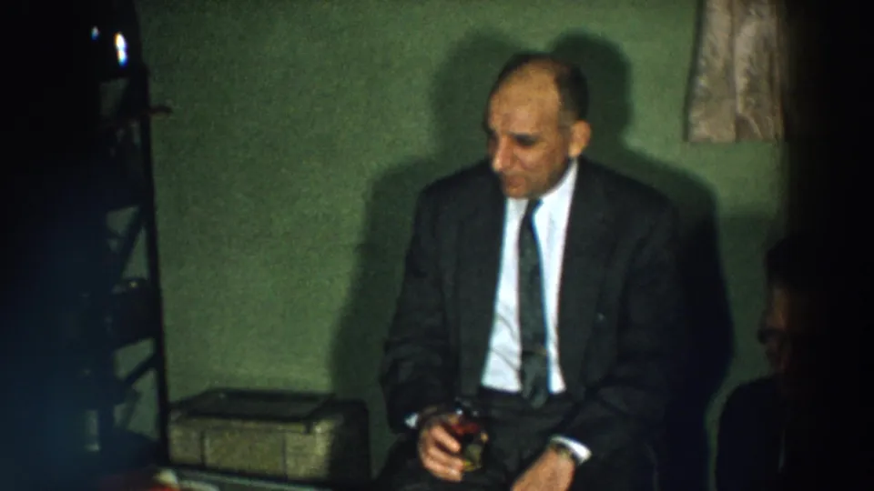 A seated party guest in 1957 Wheeling, Ohio holds a drink in a dimly lit private interior.