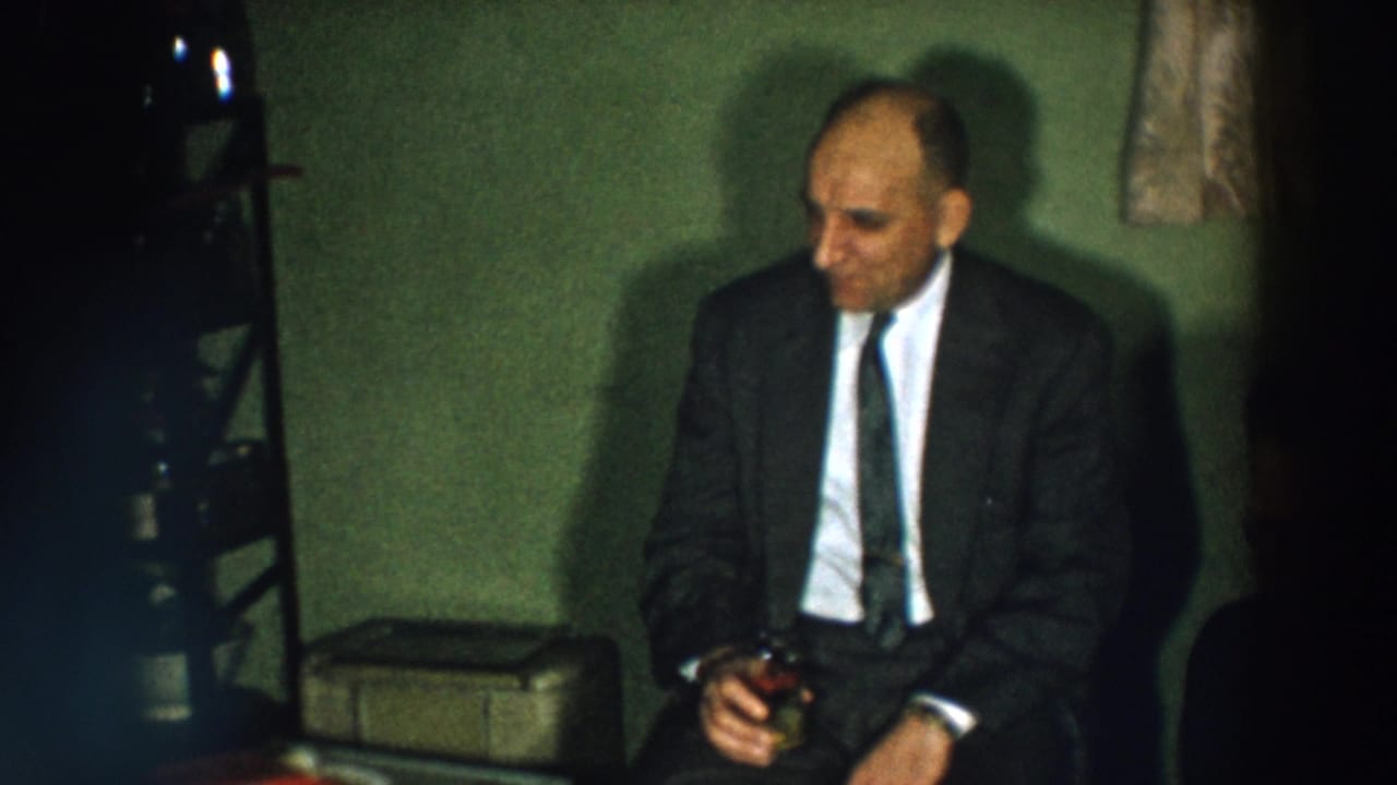 In a 1957 Wheeling, Ohio home-movie frame, a suited man sits in a dim room holding a drink during a birthday gathering.
