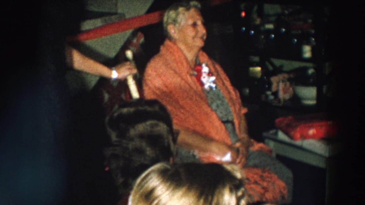 An elderly woman in a red shawl and corsage sits among guests and wrapped gifts at a 1957 Wheeling, Ohio birthday party.