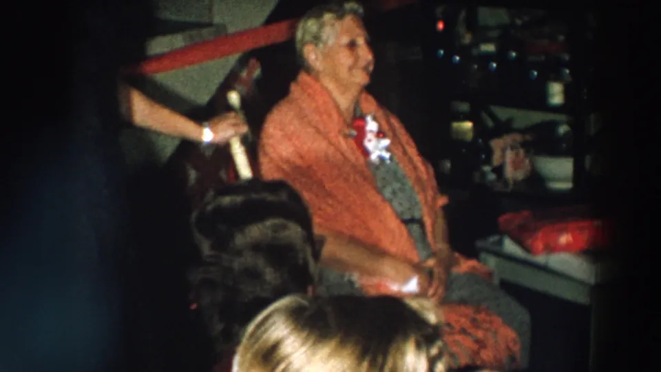An elderly woman in a red shawl and corsage sits among guests and wrapped gifts at a 1957 Wheeling, Ohio birthday party.