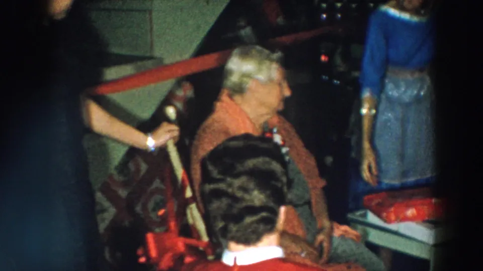The seated birthday honoree turns toward a standing guest near a wrapped present in the 1957 Wheeling, Ohio home interior.