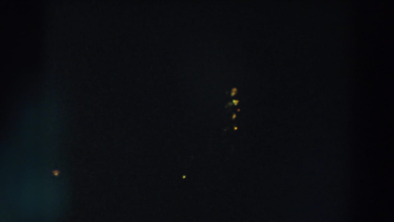 Dark night frame with a small cluster of yellow lights and a few faint points in the sky.