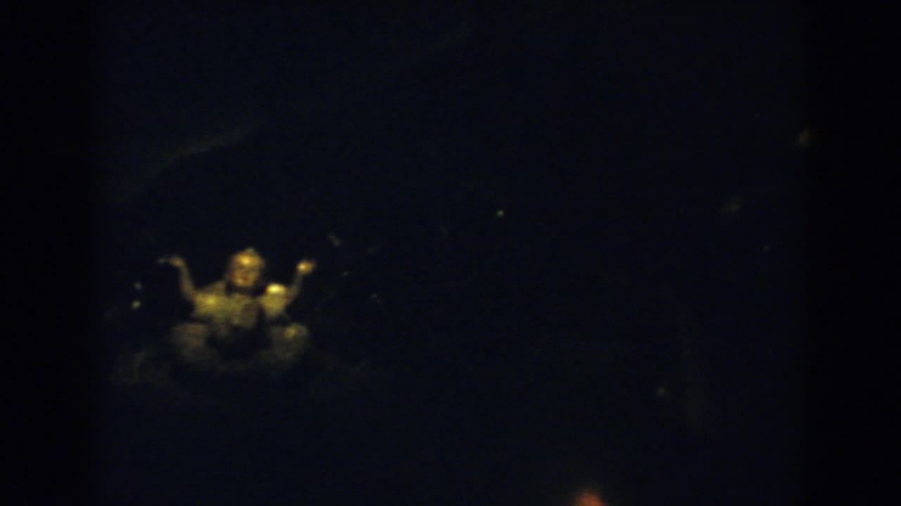 Dim frame with a warm-lit small religious figurine and a darker figure behind it.