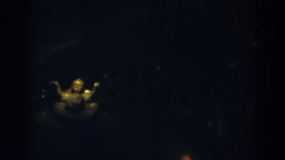 1957 8mm frame from Wheeling, Ohio, showing a warm-lit small religious figurine in a dark interior with another figure faintly behind it.