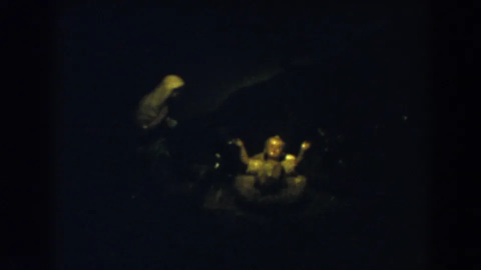 1957 8mm frame from Wheeling, Ohio, showing a small illuminated devotional object with a pale background figure in soft focus.