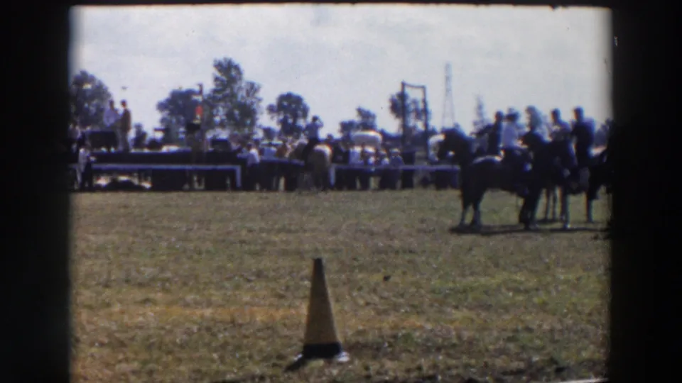 1957 Wheeling, Ohio rodeo scene with a dark horse and group at right, spectators behind a barrier, and a cone in the foreground.