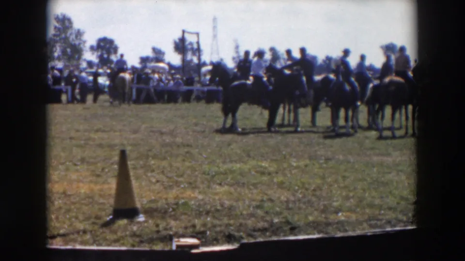 The 1957 home-movie frame shows a small line of mounted riders in a grassy ring, with a judging or spectator area behind them.