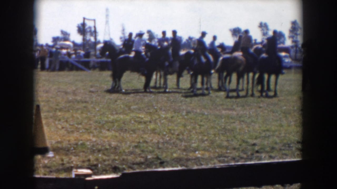 In a 1957 Wheeling, Ohio home-movie frame, men on horseback wait together on a grassy field with spectators and cars in the background.