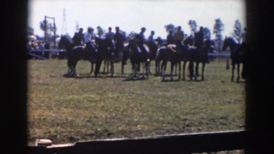 The 8mm frame shows a loose line of mounted men and horses on a grassy field, with a partial rider near the right edge and event activity behind them.