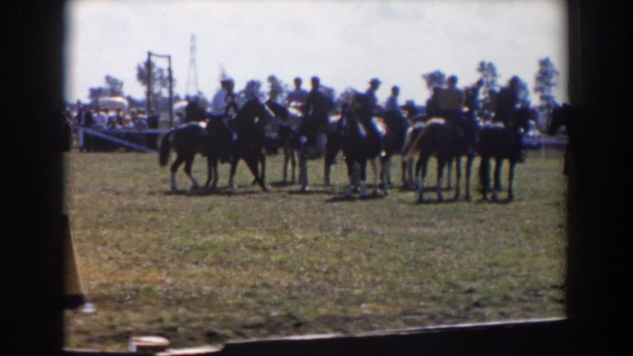 Horseback riders gather in a grassy ring with spectators and parked cars beyond the fence.