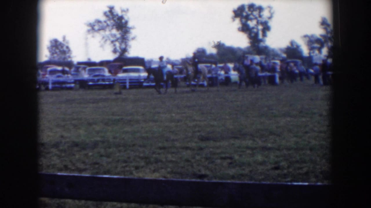 Riders move across a grassy field with spectators and rows of parked cars in the background.