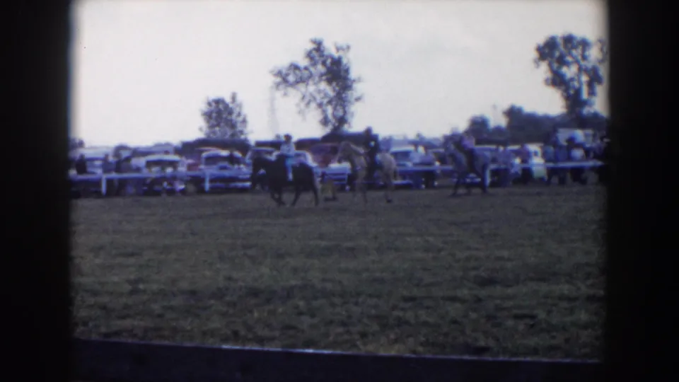 1957 Wheeling, Ohio home-movie frame showing multiple horseback riders spread across a grassy ring before spectators and parked cars.