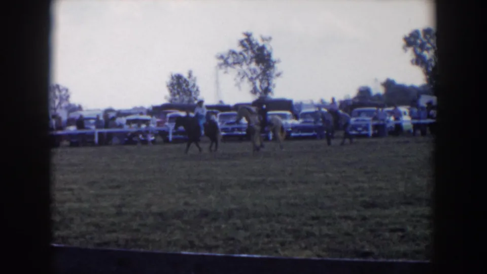 1957 Wheeling, Ohio home-movie frame showing mounted riders on grass with mid-century cars, spectators, and trees beyond a white fence.
