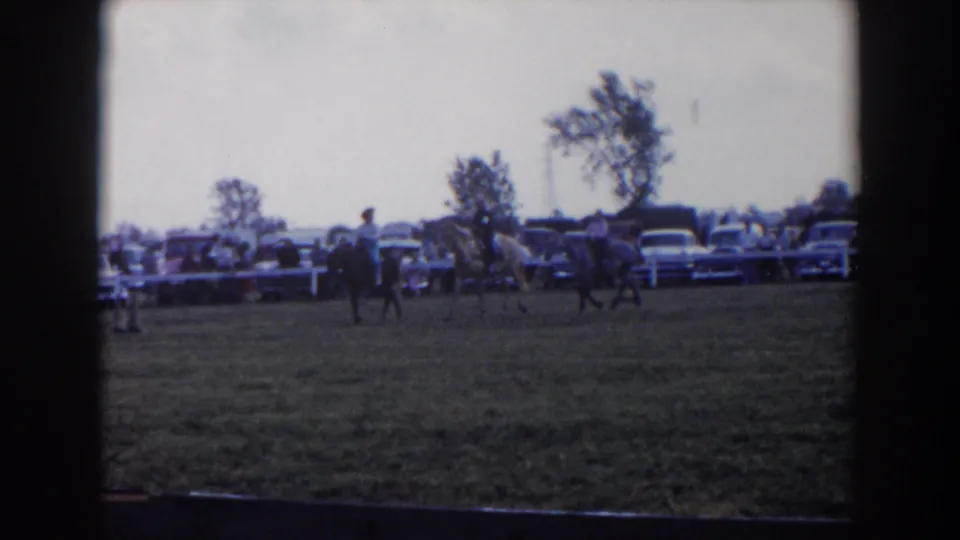 A 1957 home-movie view of horseback riders on a grassy field near a row of mid-century cars and onlookers in Wheeling, Ohio.