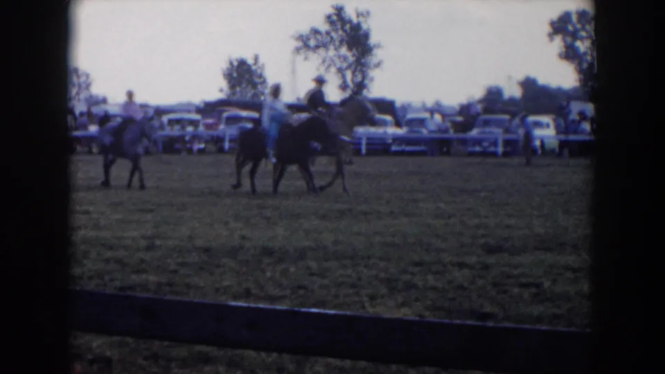 Closeer 1957 Wheeling, Ohio frame of riders on horseback crossing a field, backed by spectators, classic cars, trees, and a white fence.