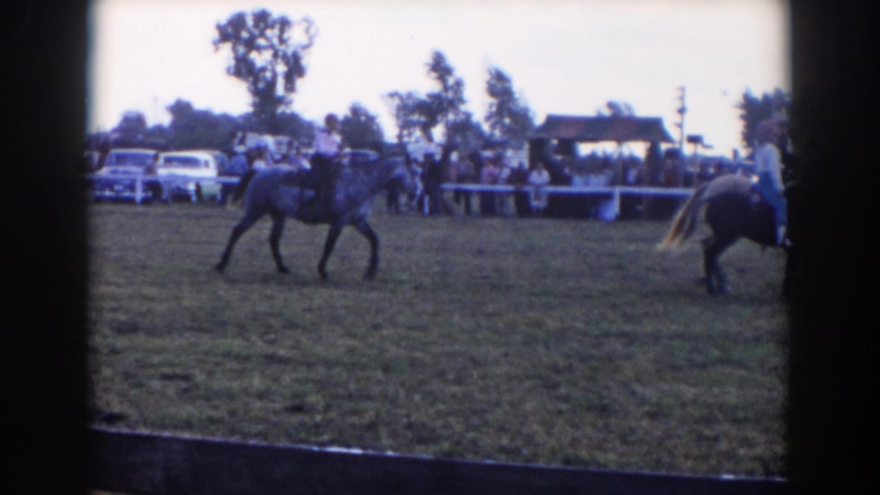 1957 Wheeling, Ohio home-movie frame showing a horseback rider in a grassy show ring with spectators and parked cars behind a fence.