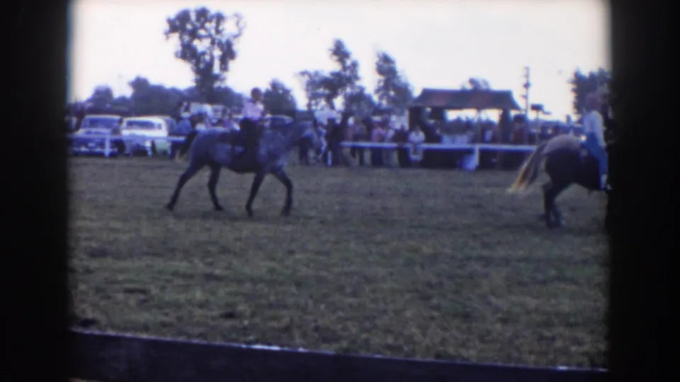 1957 Wheeling, Ohio home-movie frame showing a horseback rider in a grassy show ring with spectators and parked cars behind a fence.
