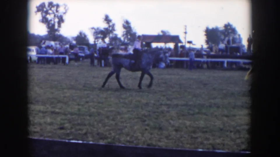 A horseback rider moves through the grassy event ring in 1957 Wheeling, Ohio, with spectators, trees, and mid-century cars behind.