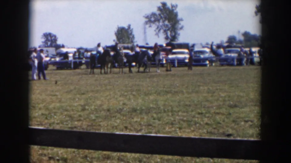 Wider home-movie view of riders, spectators, and parked 1950s cars along the edge of a grassy show area in Wheeling, Ohio.
