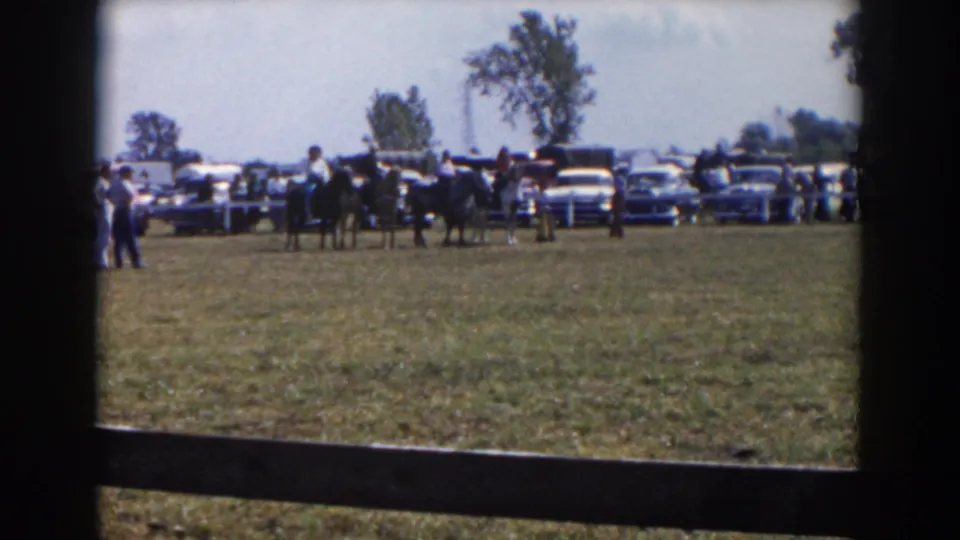 A 1957 Wheeling, Ohio fairground-style scene shows riders on horses near spectators and a row of parked mid-century cars.