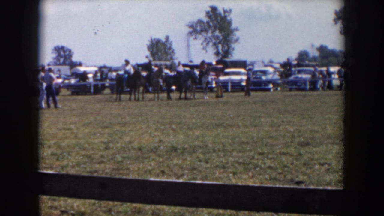 1957 Wheeling, Ohio home-movie frame showing a distant crowd, horses or dark figures, and classic cars beside an open field.