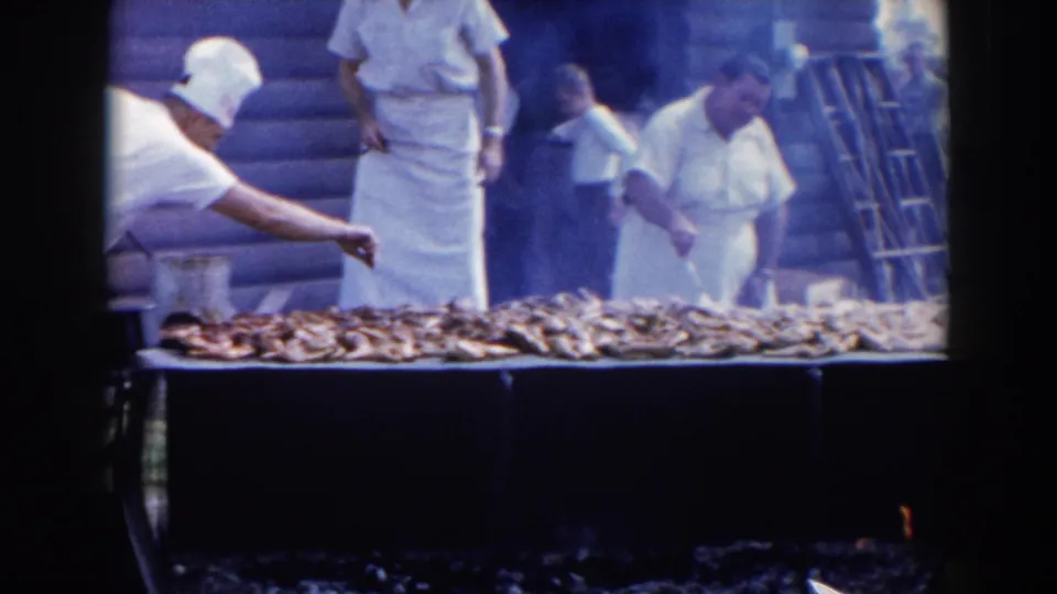 Smoke drifts around cooks in white uniforms as they tend a long grill covered with fish at the 1957 outdoor gathering.