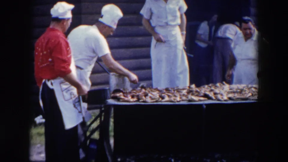 A cook in a red shirt and others in white uniforms stand around a long fish grill at the Wheeling, Ohio outdoor meal.