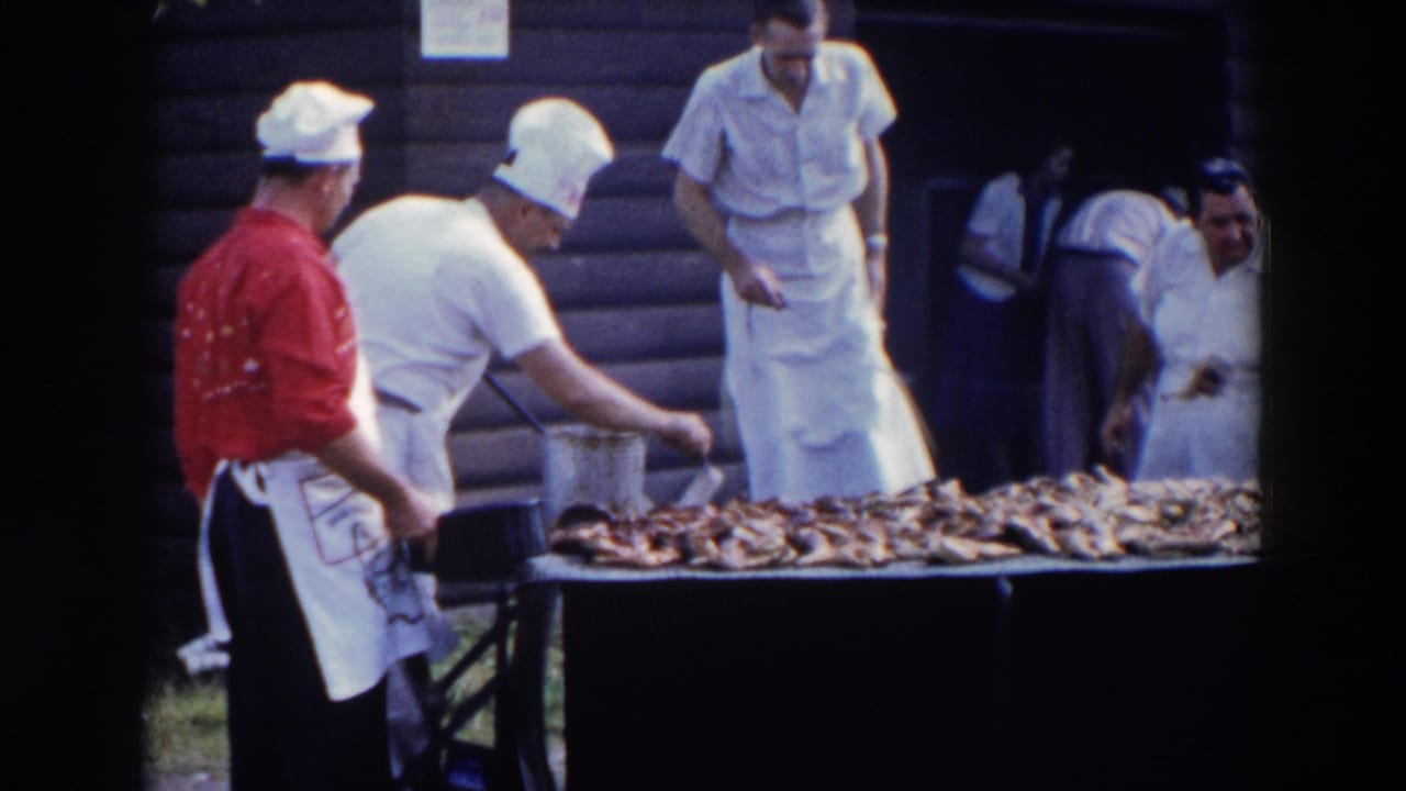 Cooks in aprons stand beside a long outdoor table covered with barbecue meat.