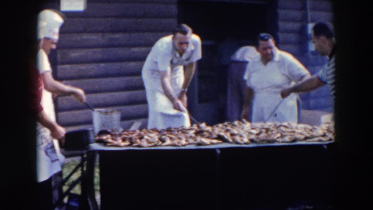 Aproned cooks use tongs near a long table piled with cooked meat outdoors.