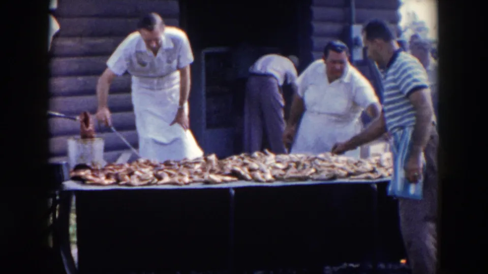 Home-movie frame of adults preparing barbecue meat outdoors, with a cook leaning over the table and a striped-shirt bystander at right.