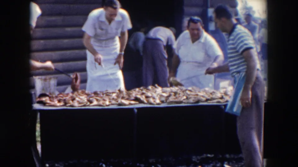 1957 Wheeling, Ohio frame showing aproned cooks, a striped-shirt man, and a long outdoor table filled with barbecue meat.