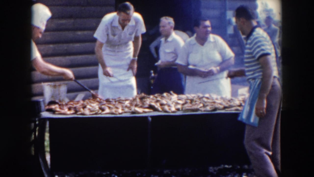 Men in aprons stand around a long grill covered with meat outside a log-sided building.