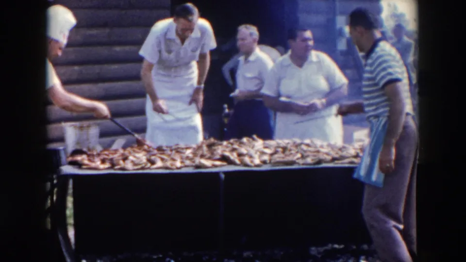 In 1957 Wheeling, Ohio, men in white aprons and chef hats tend a large outdoor grill piled with meat while bystanders stand nearby.