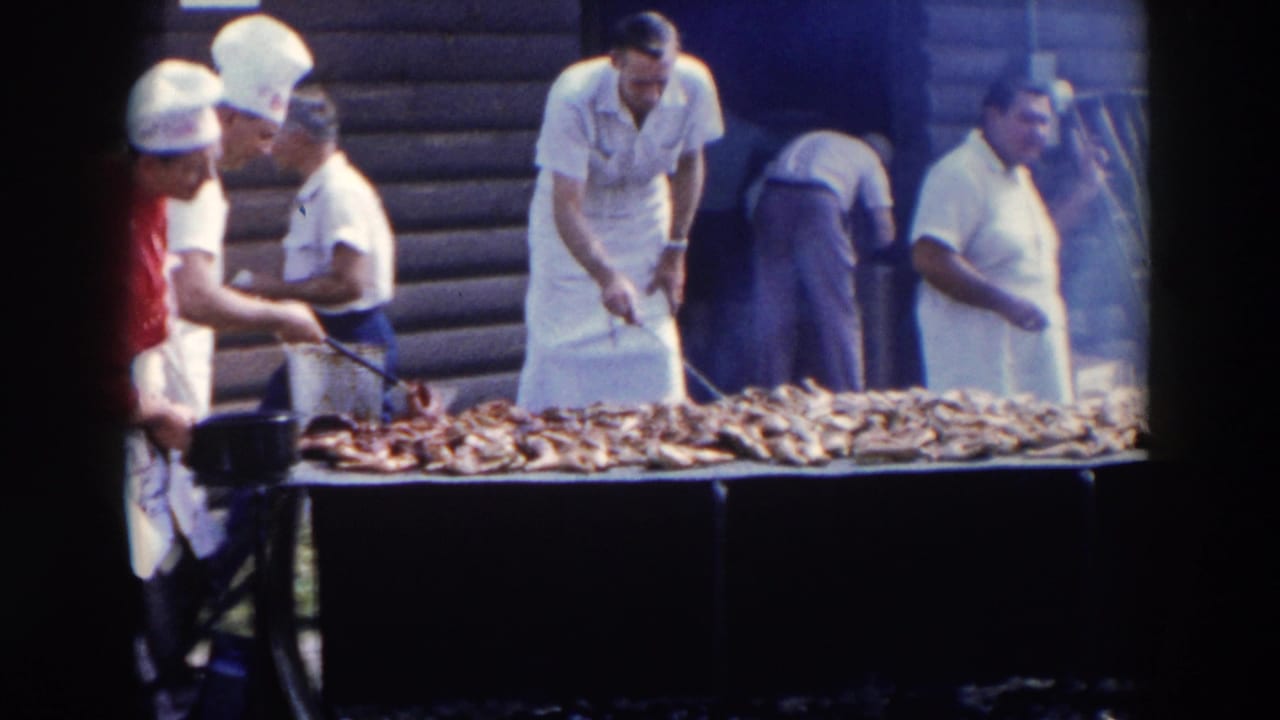 A cook in a white hat bastes meat on a long grill as other men stand near the building.