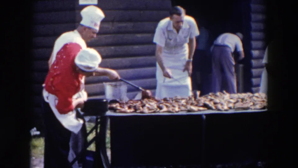 At the Wheeling cookout, a man in a red shirt and white chef hat tends meat on a long grill while another man in an apron stands behind it.