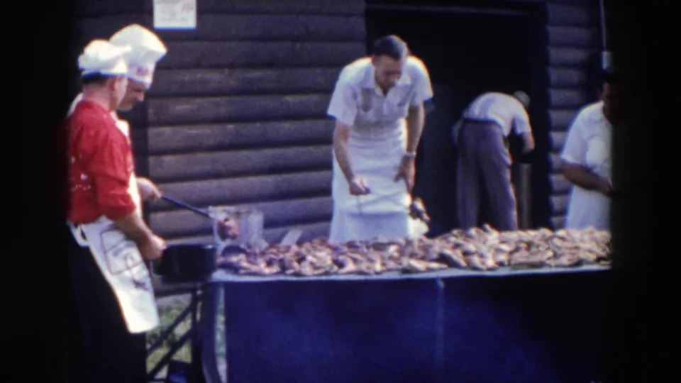 Men in aprons and chef hats continue tending a long meat-covered grill outside a log-sided building in 1957 Wheeling, Ohio.