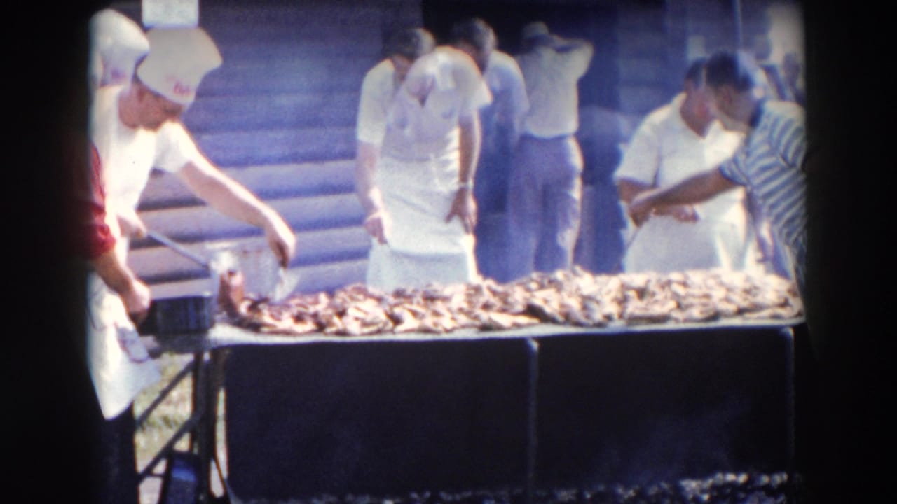 1957 Wheeling, Ohio home-movie frame showing men in white aprons working around a large barbecue grill piled with meat.