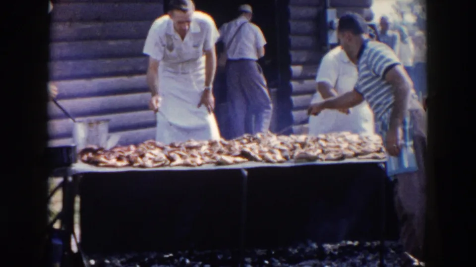 Men in aprons and casual shirts gather around a large outdoor barbecue grill in 1957 Wheeling, Ohio footage.