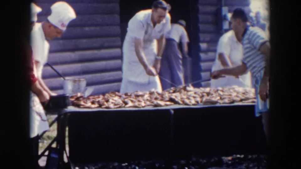 Home-movie frame from Wheeling, Ohio showing aproned men using long-handled tools over a crowded outdoor barbecue grill.