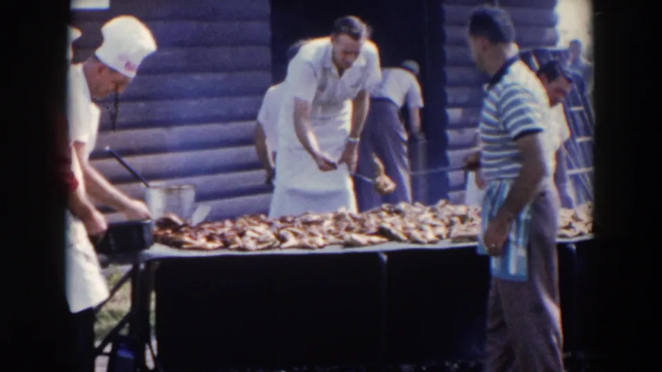1957 8mm frame showing men beside a large outdoor barbecue grill, with one man in an apron reaching toward the cooking meat.