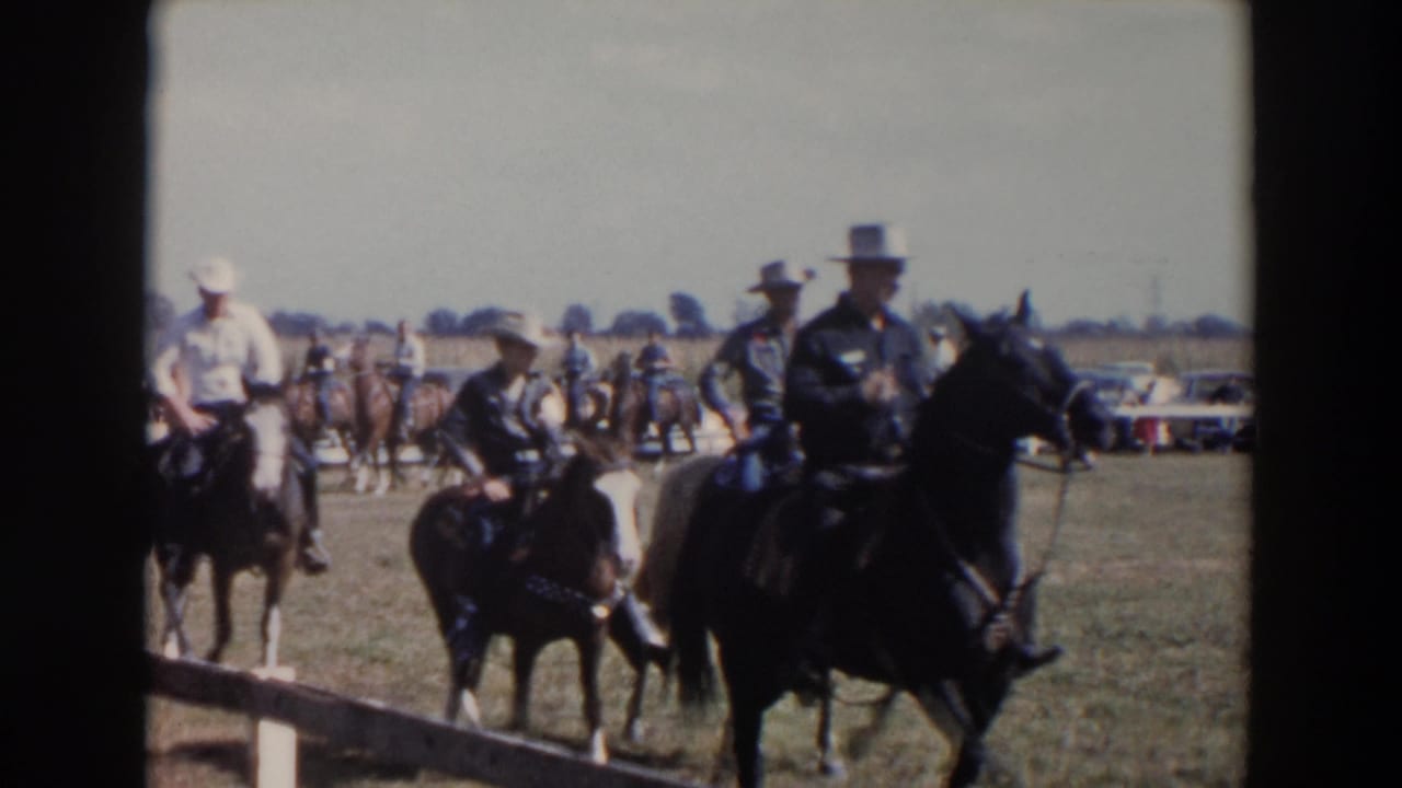 Mounted riders in hats cross a grassy fairground with spectators and cars behind them.