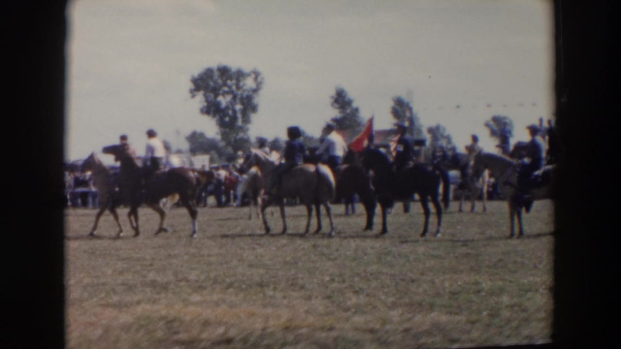 Horse riders gather on a field near spectators, trees, parked cars, and a flag.
