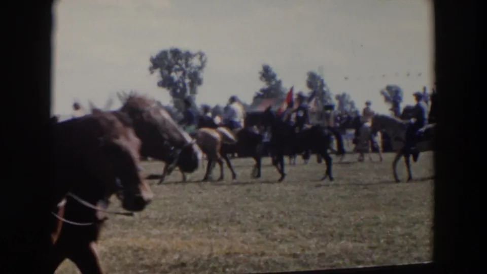 Close foreground horses partially frame the 1957 fairground scene, with mounted riders and spectators visible farther back across the grassy field.