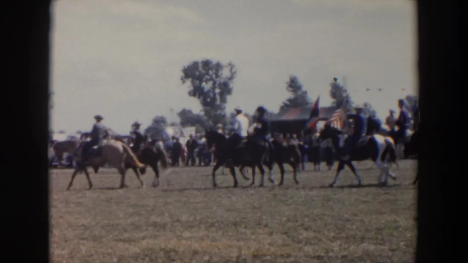 Wider 1957 Wheeling, Ohio rodeo parade view with multiple horse riders, spectators, American flags, and a small tent or stand on the field.